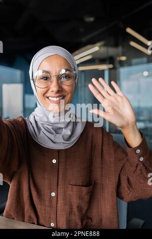 Close-up photo. Vertical photo. A young female blogger in a hijab talks to the camera, holds a phone in her hand. Films a blog, a podcast. He greets the camera with his hand, smiles. Stock Photo