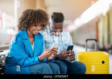 Black Couple Playing Games On Smartphones While Waiting Train At Railway Station Stock Photo