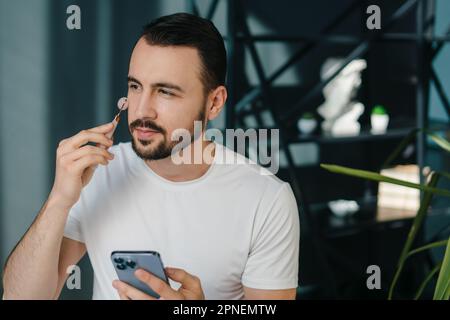 Bearded man using jade face roller for beauty facial massage therapy at home. Facial roller for massage over the skin. Stock Photo