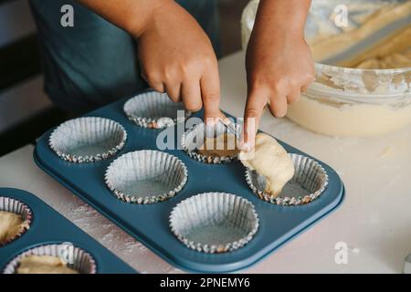 Close-up view of baby girl's hands holding spoon pouring cake batter in cup cake moulds. Children helping parents. Stock Photo