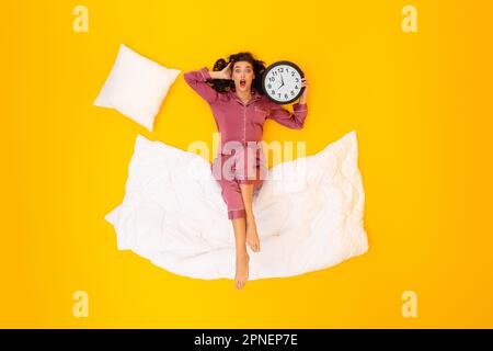 Shocked Lady Holding Alarm Clock Lying On Yellow Background Stock Photo
