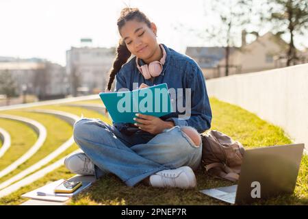 Remote study concept. Black teen lady with laptop preparing for exam outdoors and taking notes, sitting on lawn Stock Photo