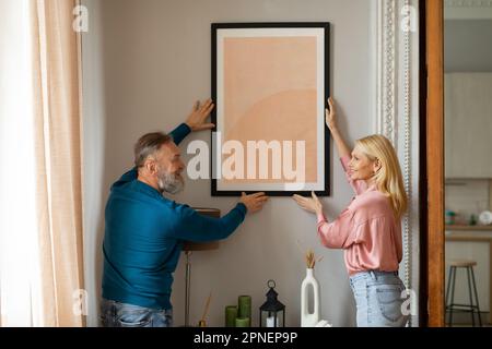 Happy Mature Couple Hanging Picture In Frame On Wall Indoors Stock Photo