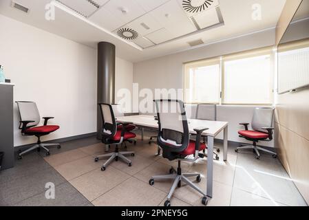 An office with several swivel chairs with a red seat, a wooden table, windows with blinds and technical ceilings Stock Photo