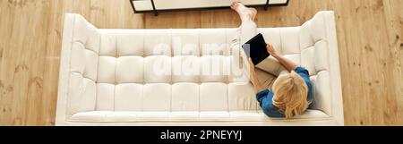 Top view of mature woman in casual wear using tablet pc while working from home, doing some paperwork, sitting on a couch in modern apartment Stock Photo