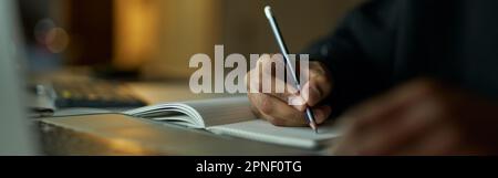 Writing ideas. Close up shot of hands of male student making notes in his notebook while studying, using laptop late at night Stock Photo