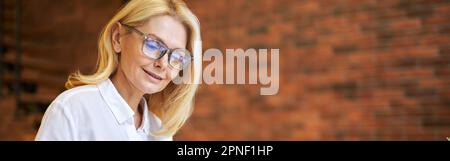 Attractive mature businesswoman in glasses using laptop and doing some paperwork at the office Stock Photo