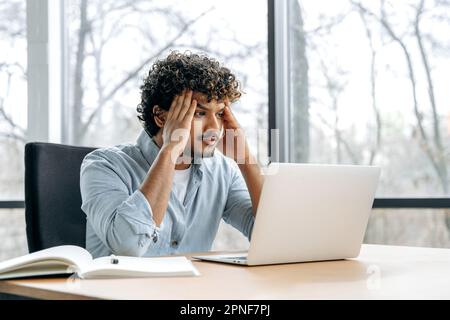 Sad worried indian or arabian curly man, financial manager, company ceo, sitting at a desk in the office, looking at a laptop screen in confusion, has work problems, experiencing anxiety, stress Stock Photo