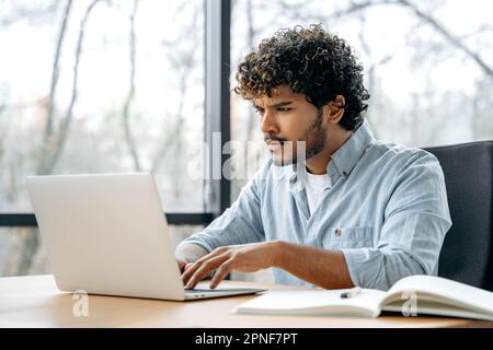 Puzzled frustrated indian or arabian curly man, financial manager, company ceo, sitting at a desk in the office, looking at a laptop screen in confusion, experiencing misunderstanding, anxiety, stress Stock Photo