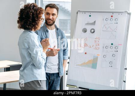 Successful positive business colleagues, caucasian man and african american woman, office coworkers, stand in the modern office near the marker board, analyze financial charts, profits, discuss risks Stock Photo