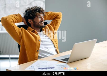 Relaxation and rest. Satisfied indian or arabian man, company employee, sits at his work desk, rests from work in a laptop, relaxes with his hands behind his head, dreaming about weekend, smiles Stock Photo