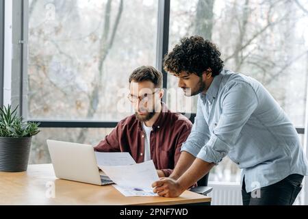 Focused busy successful business colleagues, caucasian man and indian, work together in the modern office, solve work issues, analyze results, develop a business strategy Stock Photo