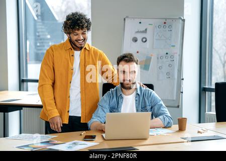 Two colleagues, Indian and Caucasian men, stylishly dressed, freelancers, designers, company employees, work in office on a project, develop a concept, plan a strategy, look at a laptop screen, smile Stock Photo