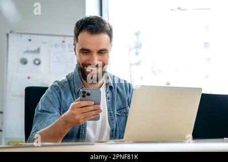 Satisfied caucasian man with beard, freelancer, programmer, seo, sits at a table in the office, uses his smartphone, messaging on social networks, conducts work correspondence, watches news, smiles Stock Photo