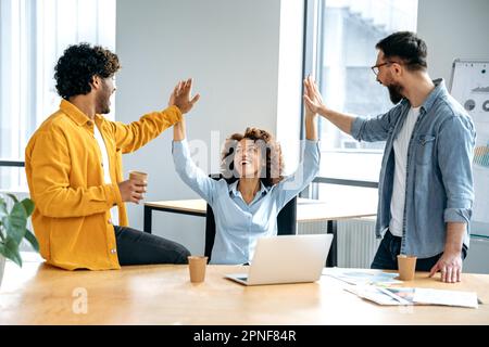 Cheerful colleagues, multiracial team, sitting in a creative office, working on a new project, a startup, developing a strategy and deadlines, happy with the result, celebrate success, achievement Stock Photo