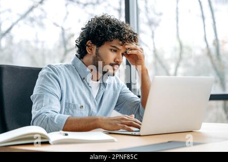 Tired sad indian or arabian businessman, company employee, manager, sitting at workplace in the office, suffering from headache, migraine, experiencing stress, anxiety, need a rest Stock Photo