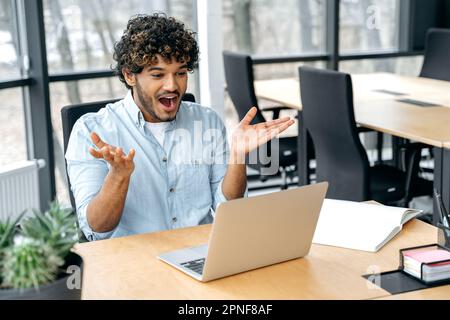 Happy excited indian or arabian businessman, company employee, manager, sitting at workplace in the office, looks in amazement at a laptop screen, received unexpected news, rejoices in success, smile Stock Photo