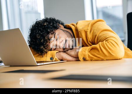 Fatigue concept. Tired exhausted indian or arabian, office employee or freelancer, fell asleep on a desk while working in a laptop in the modern office, is overworked, needs rest Stock Photo