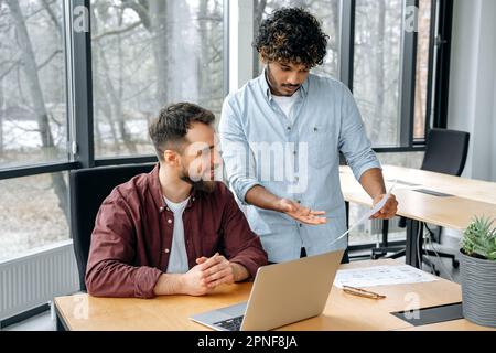 Two successful creative male business partners of different nationalities, talking at workplace with laptop in the office, brainstorming on new project, discussing ideas, planning business strategy Stock Photo