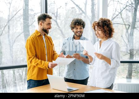 Young successful business team of creative multiracial people, gathered together in the modern office for brainstorming, analyze documents and reports, generate new ideas, share opinions, smile Stock Photo
