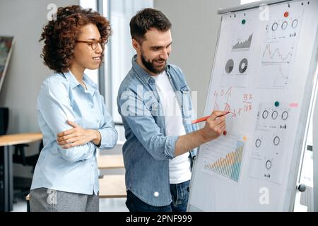 Positive successful colleagues of different nationalities, a man and a woman, office employees, stand in the modern office near the marker board, analyze financial charts, profits, discuss risks Stock Photo