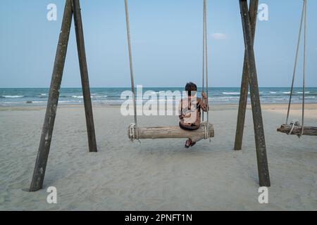 Back view of senior woman sitting on a wooden plank swing facing the sea. Retirement or travel vacation concept. Stock Photo