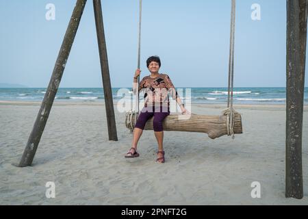 Senior Asian woman relaxing on a wood plank swing on the beach. Retirement or travel vacation concept. Stock Photo