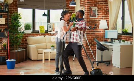 Silly couple singing song and cleaning apartment floors, using mop and vacuum cleaner at home. Diverse man and woman dancing and listening to music, washing dust and mess having fun. Stock Photo