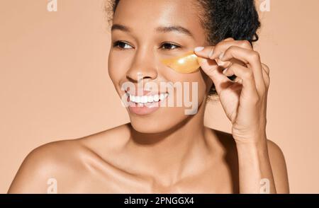 Curly swarthy woman applying under eye golden cosmetic patches, isolated. Concept of face skincare Stock Photo