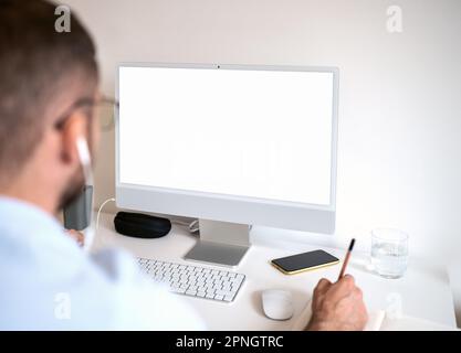 Male person sitting at desk looking at computer screen and writing notes with pencil. Stock Photo