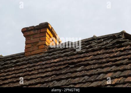 red brick chimney in the top of the village house. A roof made of tiles on the background of the sky. Stock Photo