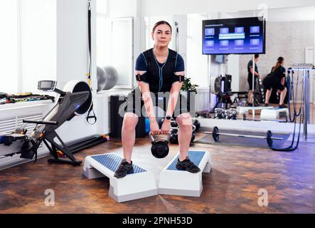 https://l450v.alamy.com/450v/2pnh53d/young-woman-in-ems-suit-standing-on-steps-and-doing-squats-with-kettlebell-female-athlete-squatting-with-weight-at-modern-gym-electrical-muscle-stim-2pnh53d.jpg