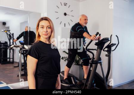 Personal female trainer with her client in ems suit in gym. Sports senior grayhaired man is exercising on elliptical trainer on background. Electrical Stock Photo