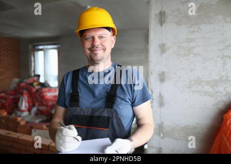 Joyful handyman, builder in uniform and hard hat looks at camera and takes notes on clipboard Stock Photo