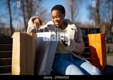 Outdoor portrait of happy black woman. She is sitting on bench in the street and looking at shopping bag. Stock Photo