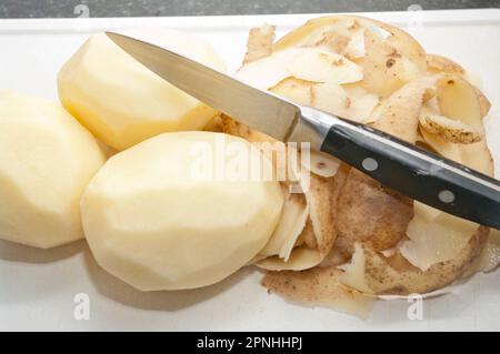 Peeled Maris Piper Potatoes with peel on a white chopping board with a potato peeling knife Stock Photo