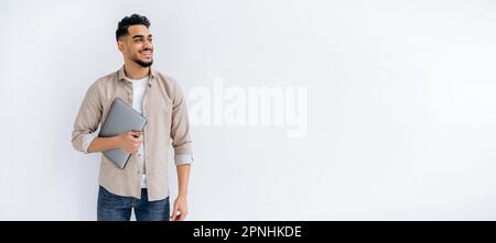 Copy-space. Panoramic photo of happy arabian or indian bearded man, in casual shirt, standing on isolated white background, holding laptop, looking to the side, smiling joyfully, dreaming, thinking Stock Photo