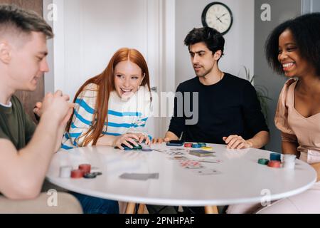 Happy overjoyed young woman laughing celebrating victory in game of poker sitting at table in living room. Group of multiethnic men and women having Stock Photo