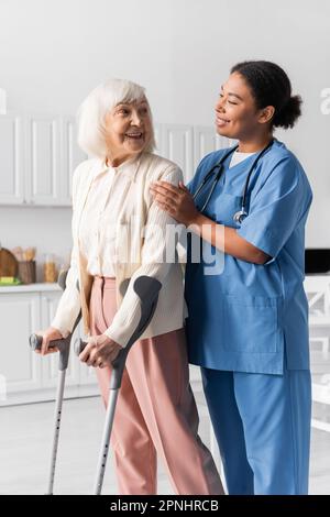 cheerful retired woman with grey hair using crutches while walking near multiracial nurse at home Stock Photo