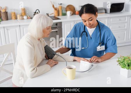 multiracial nurse measuring blood pressure of senior woman with grey hair next to cup of tea on table Stock Photo