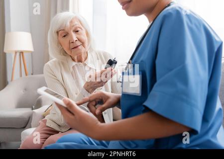 senior woman with grey hair holding glucometer near multiracial nurse using digital tablet Stock Photo