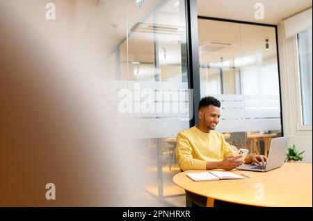 Successful freelancer. Happy millennial Indian man sitting at desk with smartphone and using laptop, smiling and typing, enjoying remote work, male student studying on the distance Stock Photo