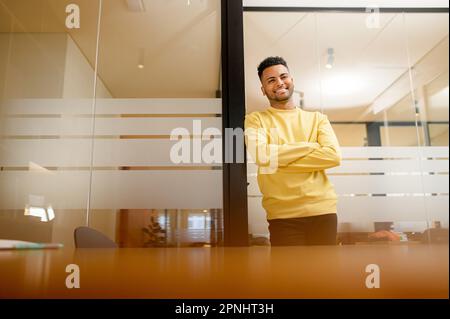 Successful confident young brunet Indian adult male entrepreneur businessman professional in casual clothes, standing with arms crossed folded at the modern office and looking at camera Stock Photo