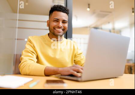 Portrait of positive indian male student sitting at the desk in front of laptop and working on a project in office, typing on the laptop, happy man looking at the camera with cheerful smile Stock Photo