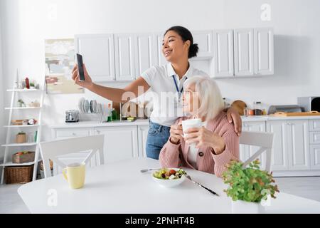 happy multiracial social worker taking selfie with retired woman during lunch in kitchen Stock Photo