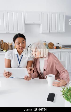 happy multiracial social worker showing digital tablet to senior woman in kitchen Stock Photo