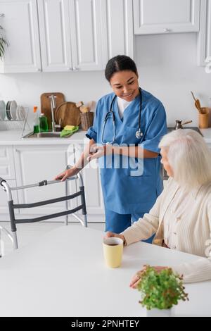 happy multiracial nurse in blue uniform pointing with hand at walker while standing near senior woman with grey hair Stock Photo