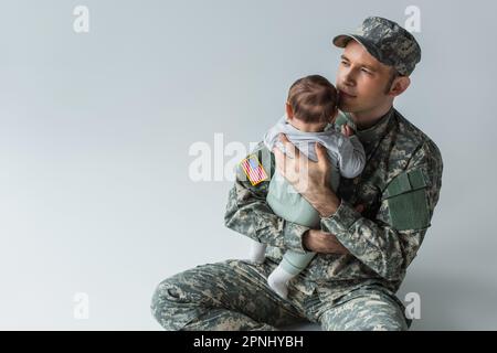 proud father in military uniform holding in arms newborn son while sitting on grey background Stock Photo