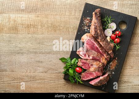 Tomahawk steak. Sliced grilled tomahawk beef steak with baked cherry tomatoes, herbs and salt on old wooden background. Preparing to grill.  Top view Stock Photo