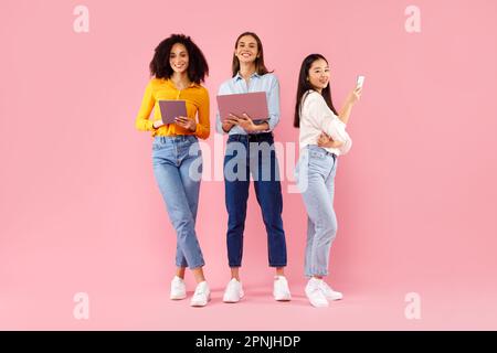 Three diverse ladies using different gadgets, networking online, standing on pink background, full length Stock Photo